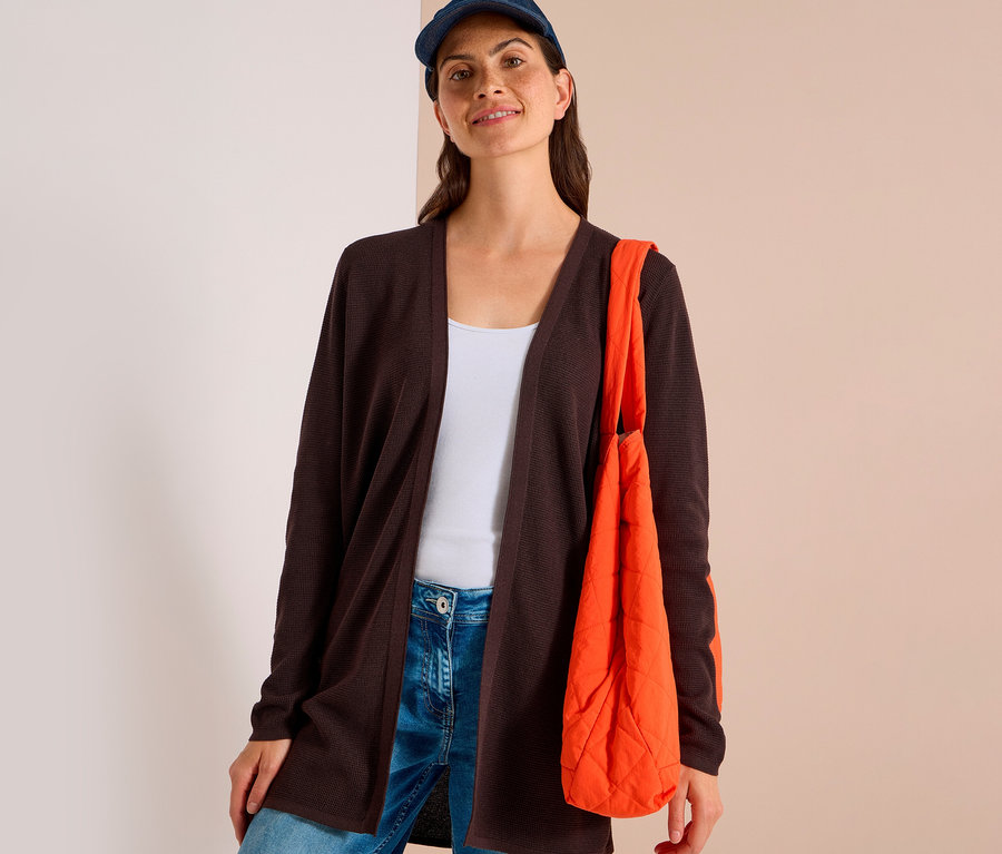 Femme portant un cardigan CECIL marron foncé sur un débardeur blanc et un jean bleu, avec une casquette bleue et un sac matelassé orange.