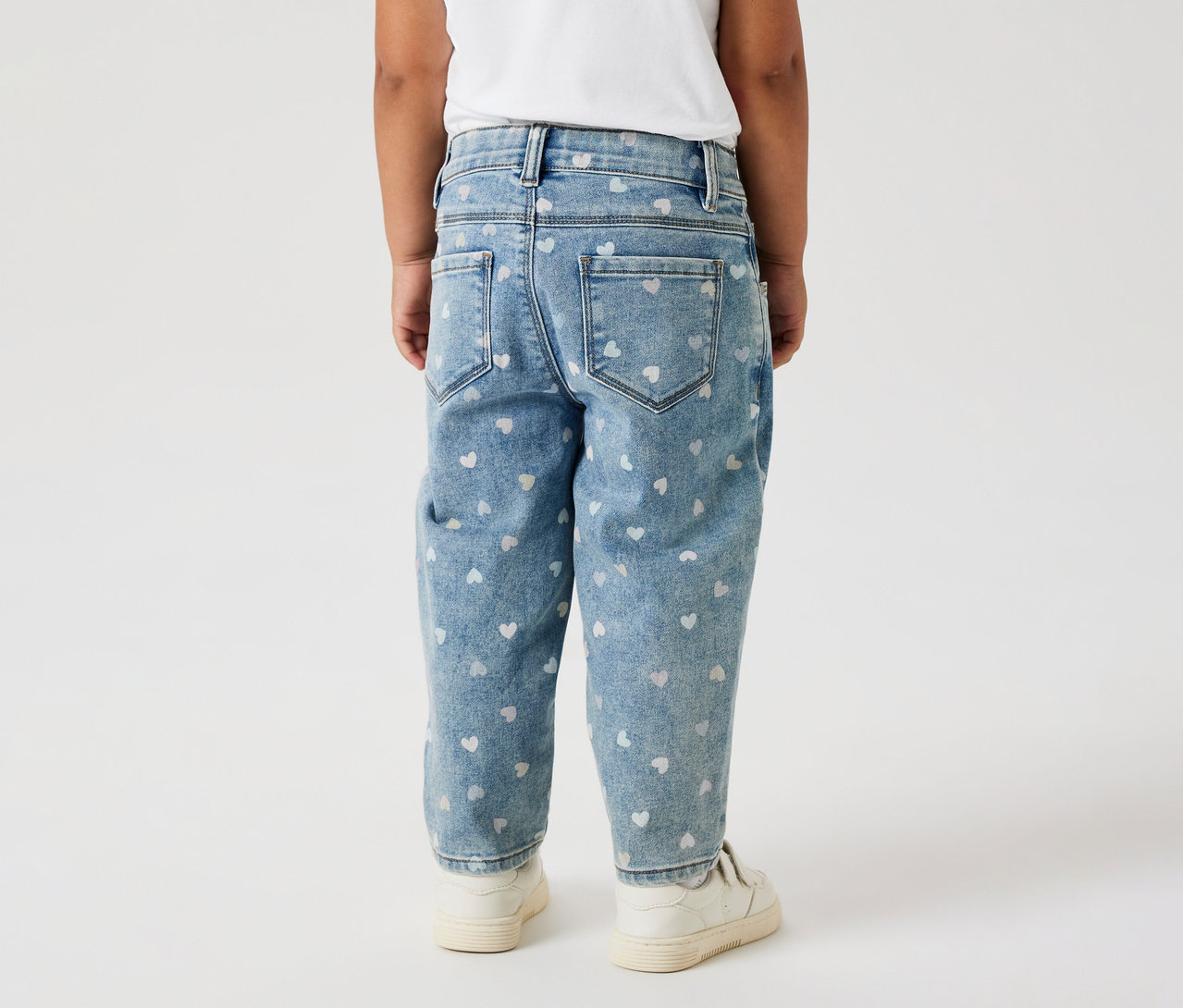 Enfant de dos, portant un jean bleu clair à motifs de cœurs blancs et des baskets blanches.
