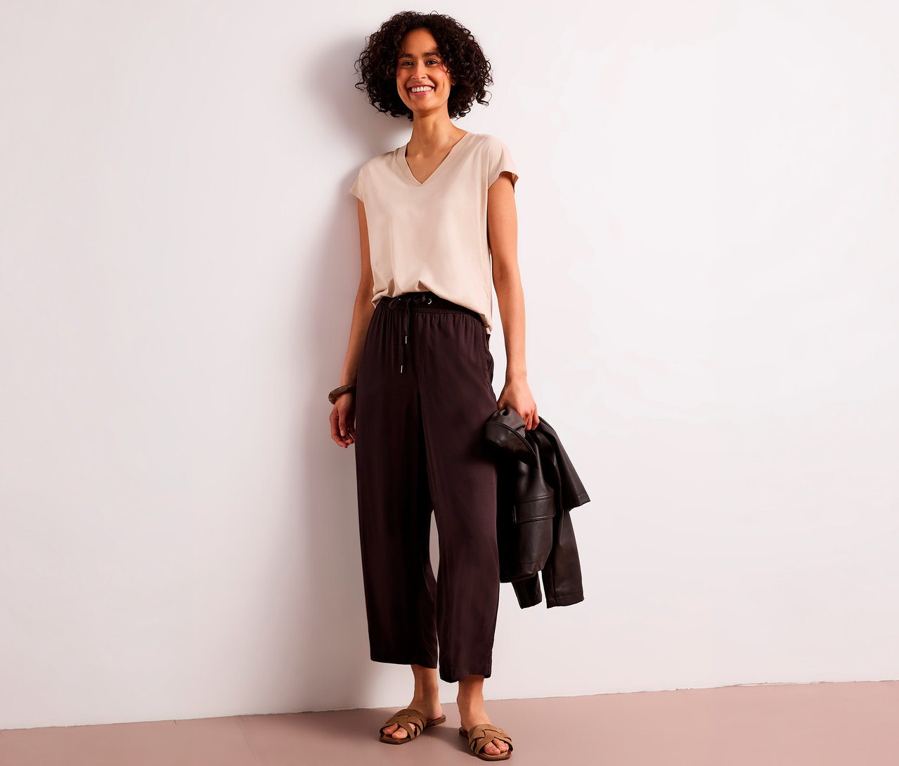 Femme souriante aux cheveux bouclés portant un top beige à col en V et un culotte 7/8 Street One marron, tenant une veste, adossée à un mur blanc.
