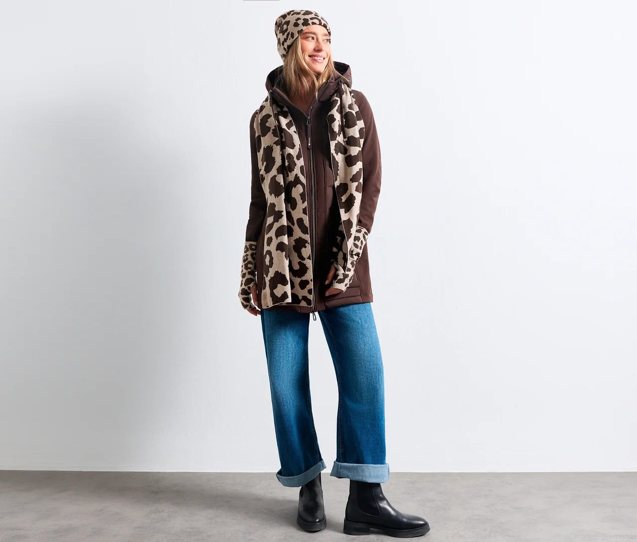 Une femme souriante porte une veste à capuche marron, un jean bleu, des bottes noires et un bonnet en tricot Street One à imprimé léopard, assorti à l'écharpe et aux mitaines.