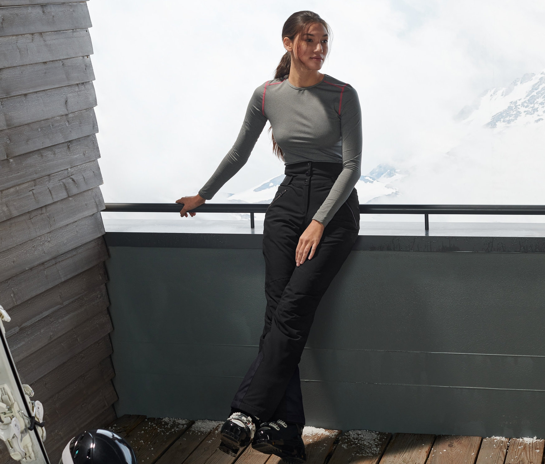Femme appuyée sur une balustrade portant un pantalon de ski taille haute noir. Montagnes enneigées en arrière-plan.