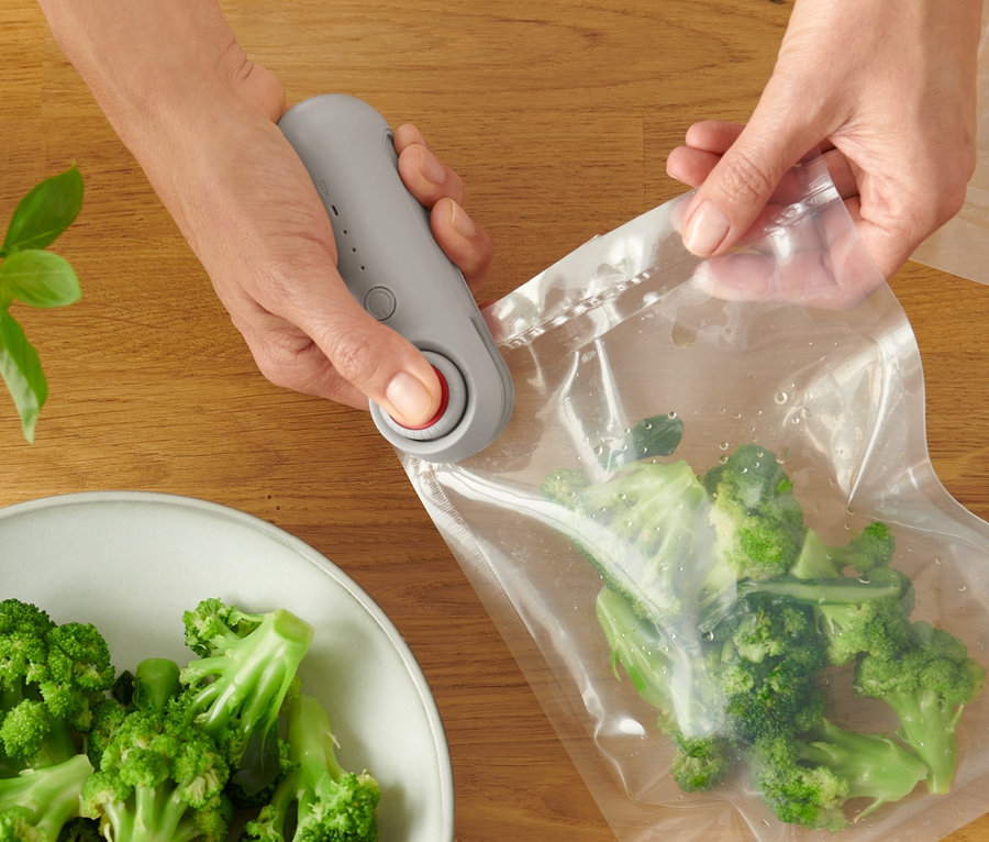 Une femme utilise une mini machine de soudage à transparents pour fermer un sac de brocolis. Du saumon, un citron et d'autres brocolis sont sur la table.