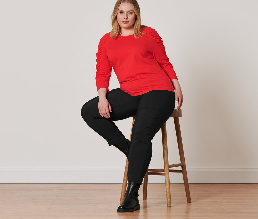 Une femme est assise sur un tabouret et porte des bottines en cuir, un pantalon en bengaline noir et un t-shirt manches 3/4 rouge.