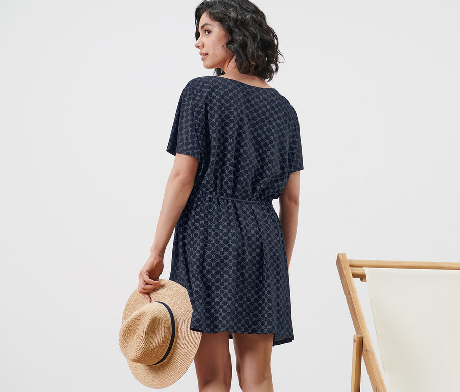 Femme en tunique de plage tenant un chapeau à côté d'une chaise longue.