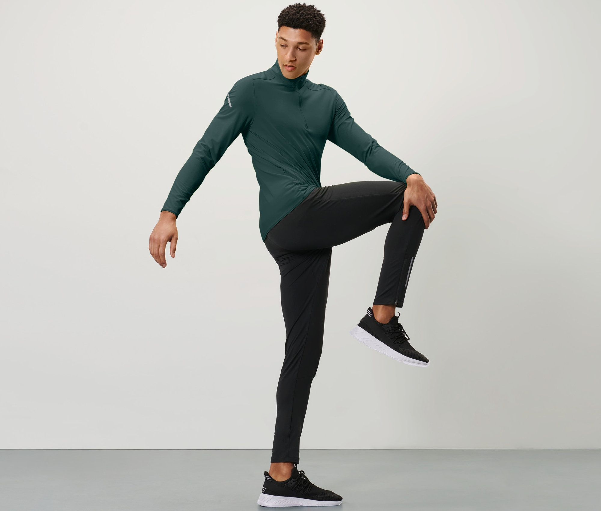 Un homme portant un haut de sport vert et un legging de course coupe-vent noir tient un genou avec une main devant sa poitrine.