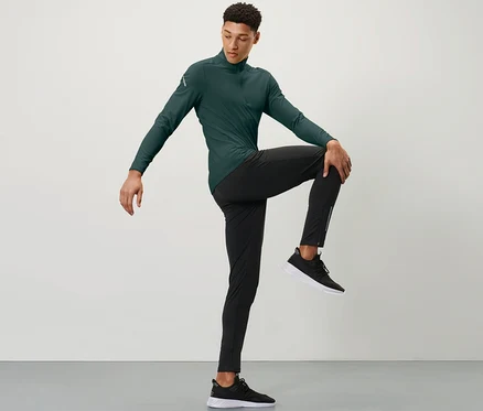 Un homme portant un haut de sport vert et un legging de course coupe-vent noir tient un genou avec une main devant sa poitrine.