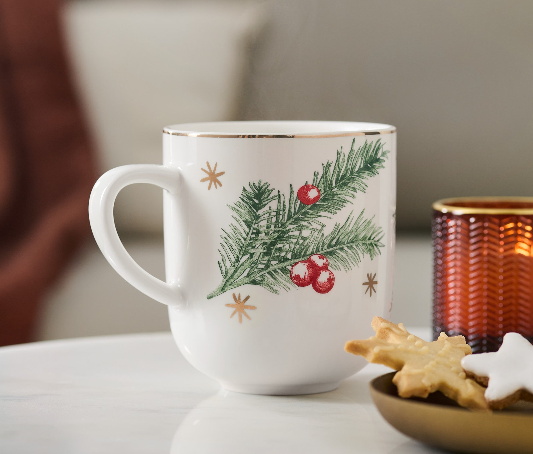 Gros plan sur un grand mug à décoration dorée et brillante avec un motif de Noël, placé à côté d'une assiette de biscuits et d'une bougie.