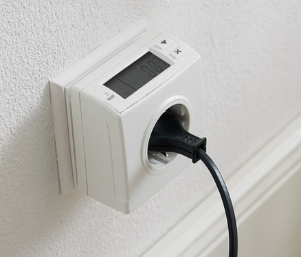 Sur un mur blanc se trouve une prise avec un appareil de mesure de la consommation d'énergie et un cordon noir branché.