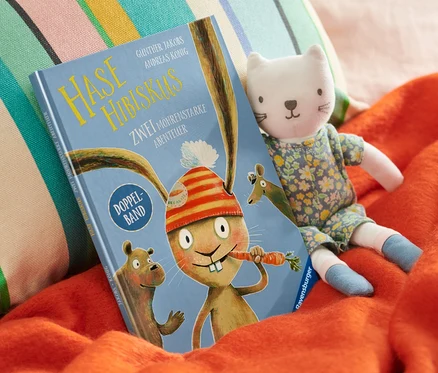 Sur une couverture orange sont posés un livre « Hase Hibiskus », une peluche chat et un coussin coloré.