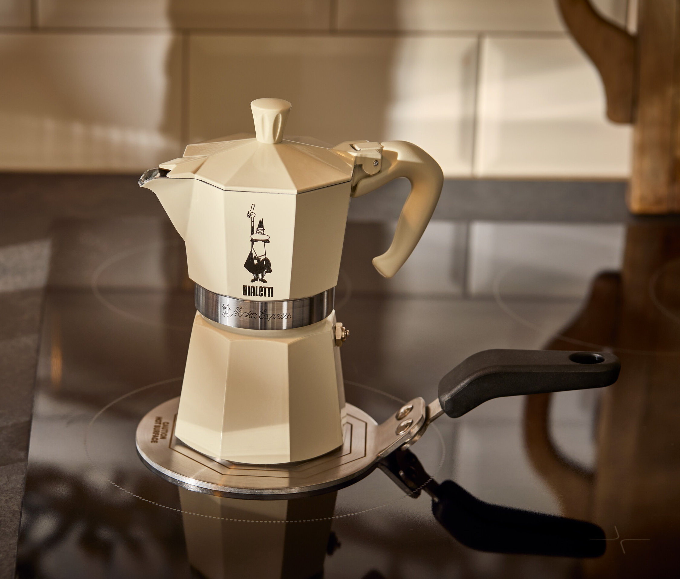 Une cafetière à expresso crème Bialetti Moka Express 190 ml est posée sur une plaque d’adaptation à induction Bialetti 13 cm à côté d’un verre d’expresso.