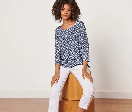 Femme assise sur un tabouret en bois portant une blouse imprimée et un pantalon blanc.