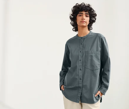 Une femme aux cheveux bouclés foncés pose sur un fond clair, portant une blouse en lin bleu-gris NAH/STUDIO.