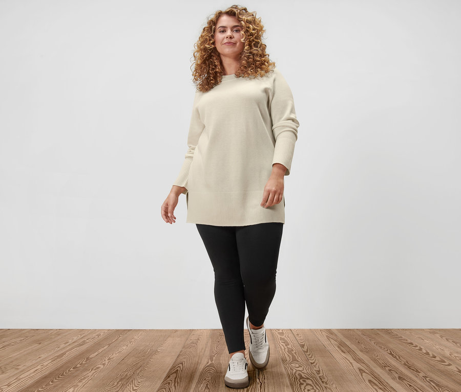 Une femme pose, portant un long pull en maille blanc crème, un legging Heavy et des baskets en cuir.