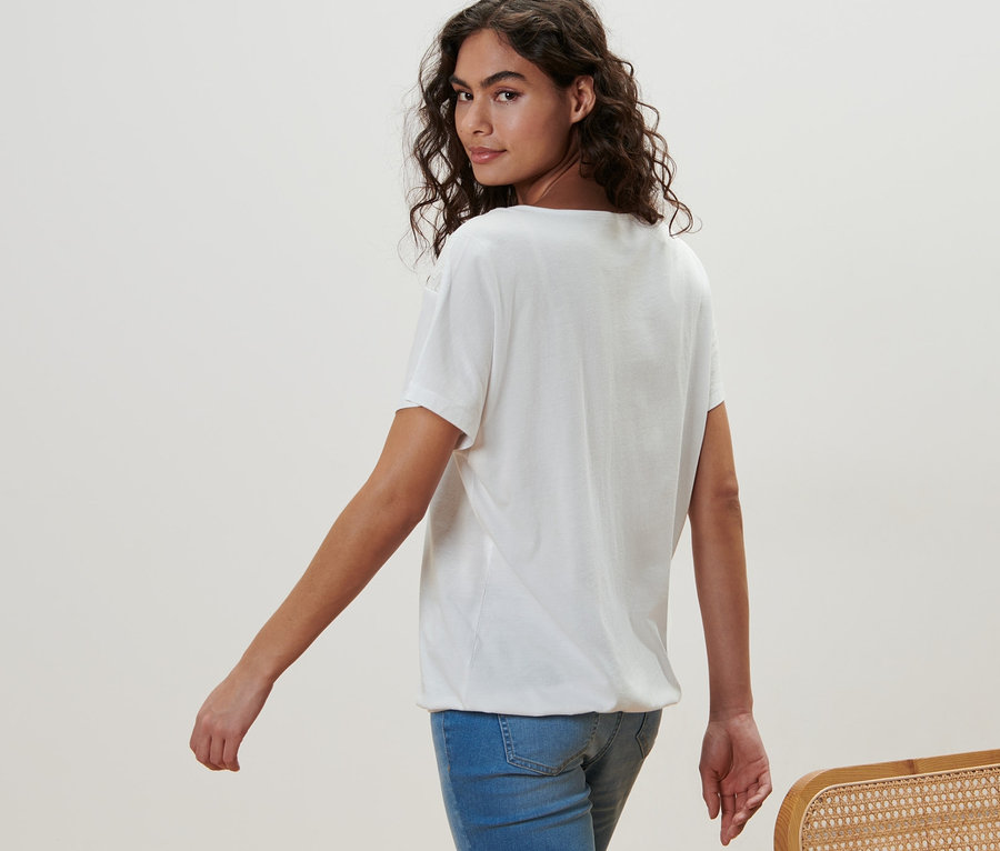 Femme marchant et regardant par-dessus son épaule. Elle porte une blouse blanche à broderie et un jean.