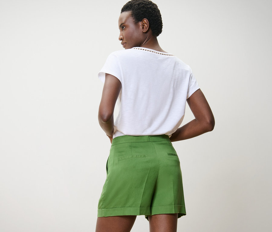 Femme à la peau foncée posant de dos portant un short en lin vert et un t-shirt blanc.