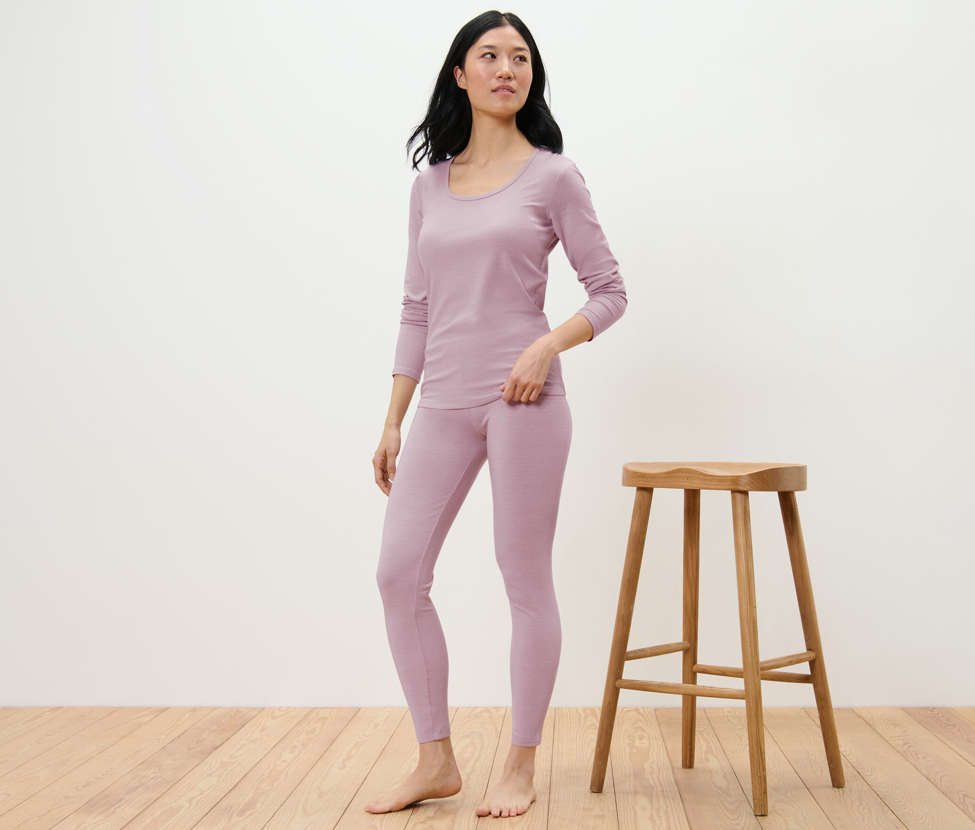 Une femme pose en leggings en laine certifiée RWS et haut à manches longues lilas à côté d'un tabouret en bois.