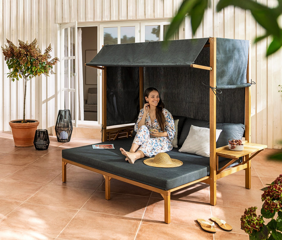 Une femme assise dans une chaise de plage Lounge « Tinus » avec repose-pieds extensible. À l'arrière-plan, une table à rallonge « Elin » et un fauteuil pour table à manger avec tressage textile.