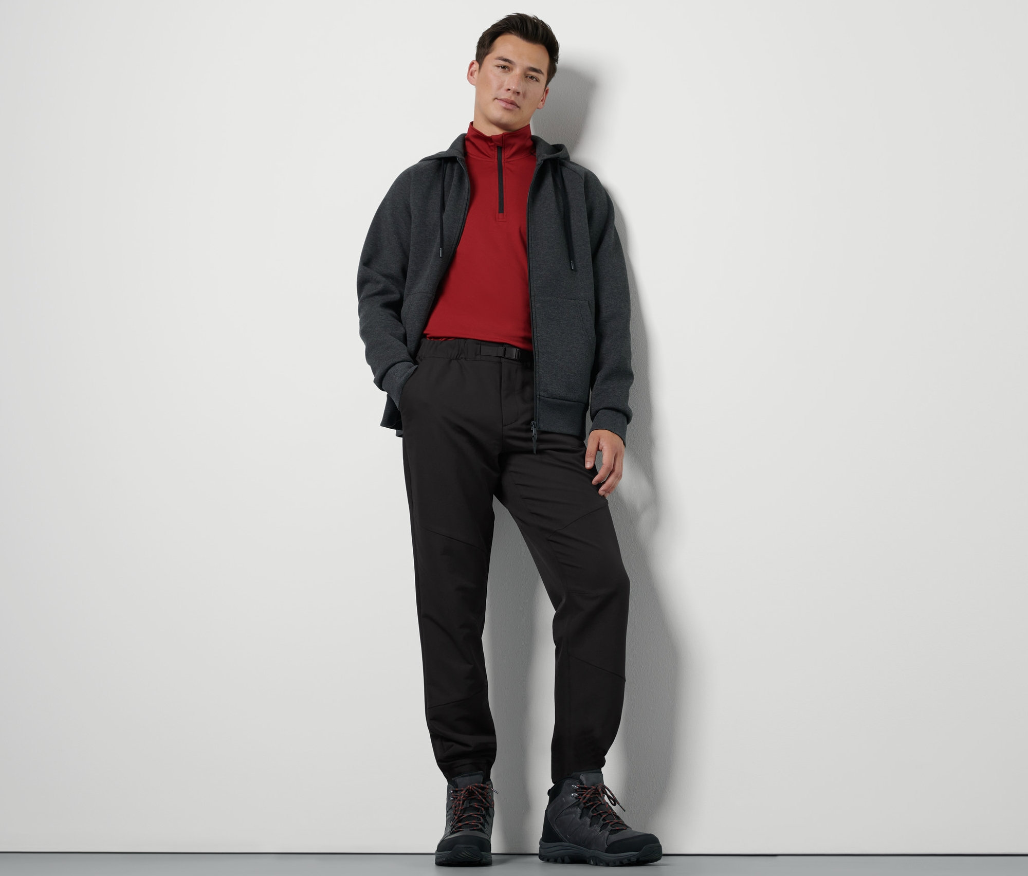 Un homme debout porte un pantalon thermique avec ceinture, un sweat à fermeture éclair et des chaussures de randonnée.