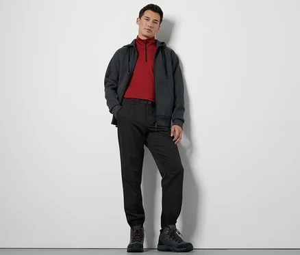Un homme debout porte un pantalon thermique avec ceinture, un sweat à fermeture éclair et des chaussures de randonnée.