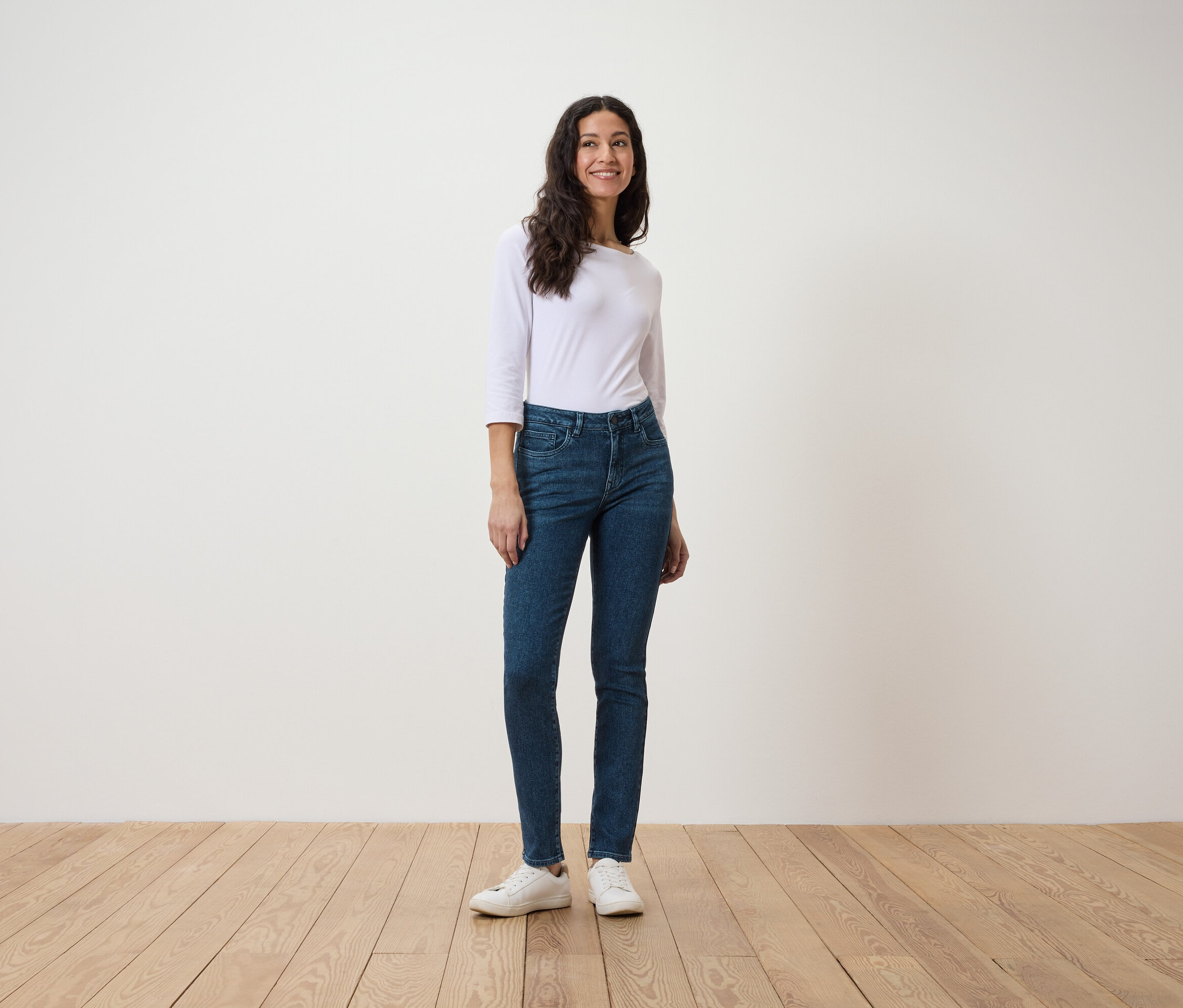 Une femme se tient sur un plancher en bois, portant une chemise blanche à manches trois-quarts et un jean bleu.