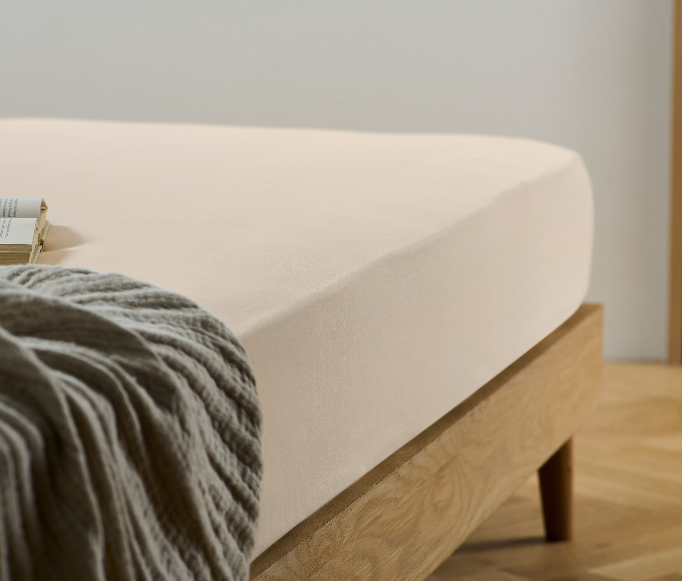 Un drap-housse beige sur un lit avec un cadre en bois. Un livre et une couverture grise sont posés sur le lit.
