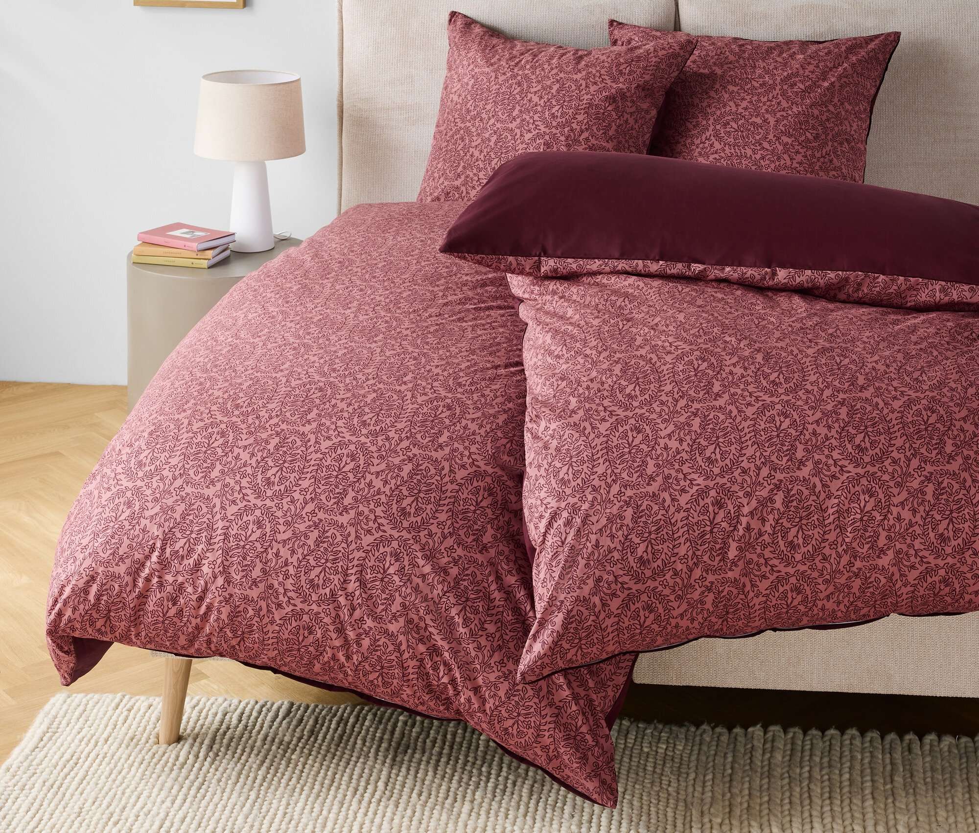 Linge de lit Ă motif floral dans les tons rose et bordeaux sur un lit avec une tĂȘte de lit beige.