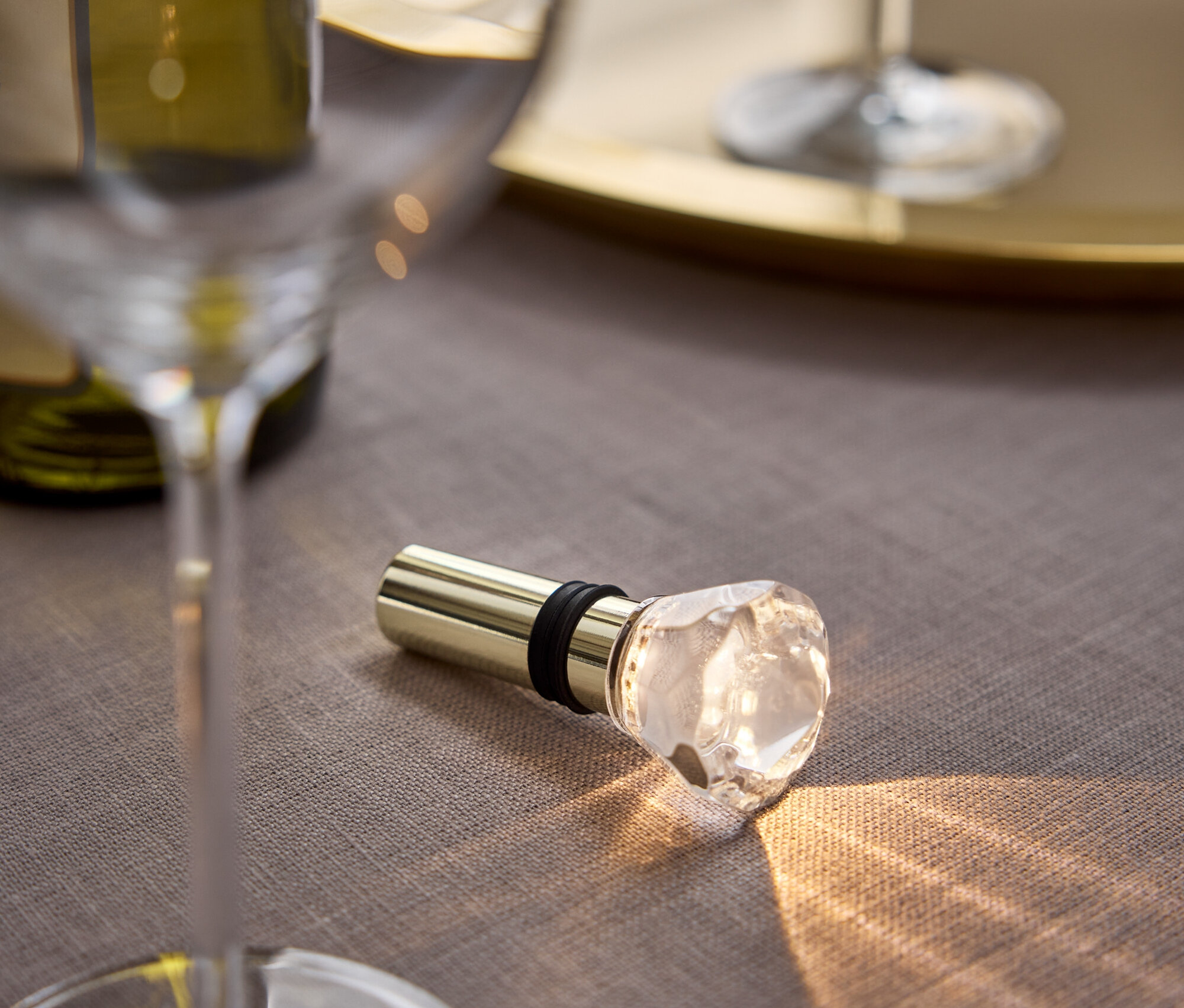 Sur une nappe grise se trouvent un bouchon de vin doré avec une tête en cristal, à côté d'un verre et d'une bouteille de vin.
