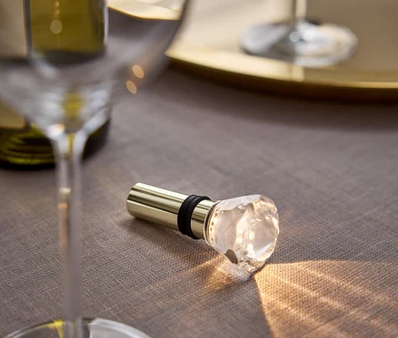 Sur une nappe grise se trouvent un bouchon de vin doré avec une tête en cristal, à côté d'un verre et d'une bouteille de vin.