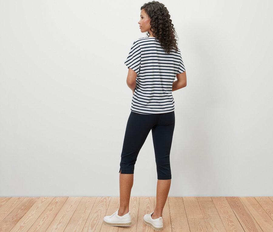 Une femme se tient dos à la caméra et porte un chemisier à encolure dégagée à rayures, un pantacourt bleu marine et des baskets blanches.