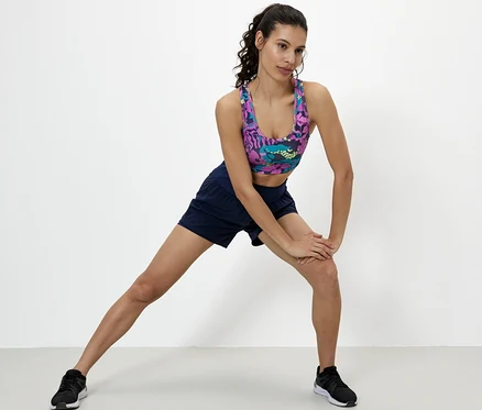 Femme étire sa jambe, porte une brassière de sport, un short et des chaussures.