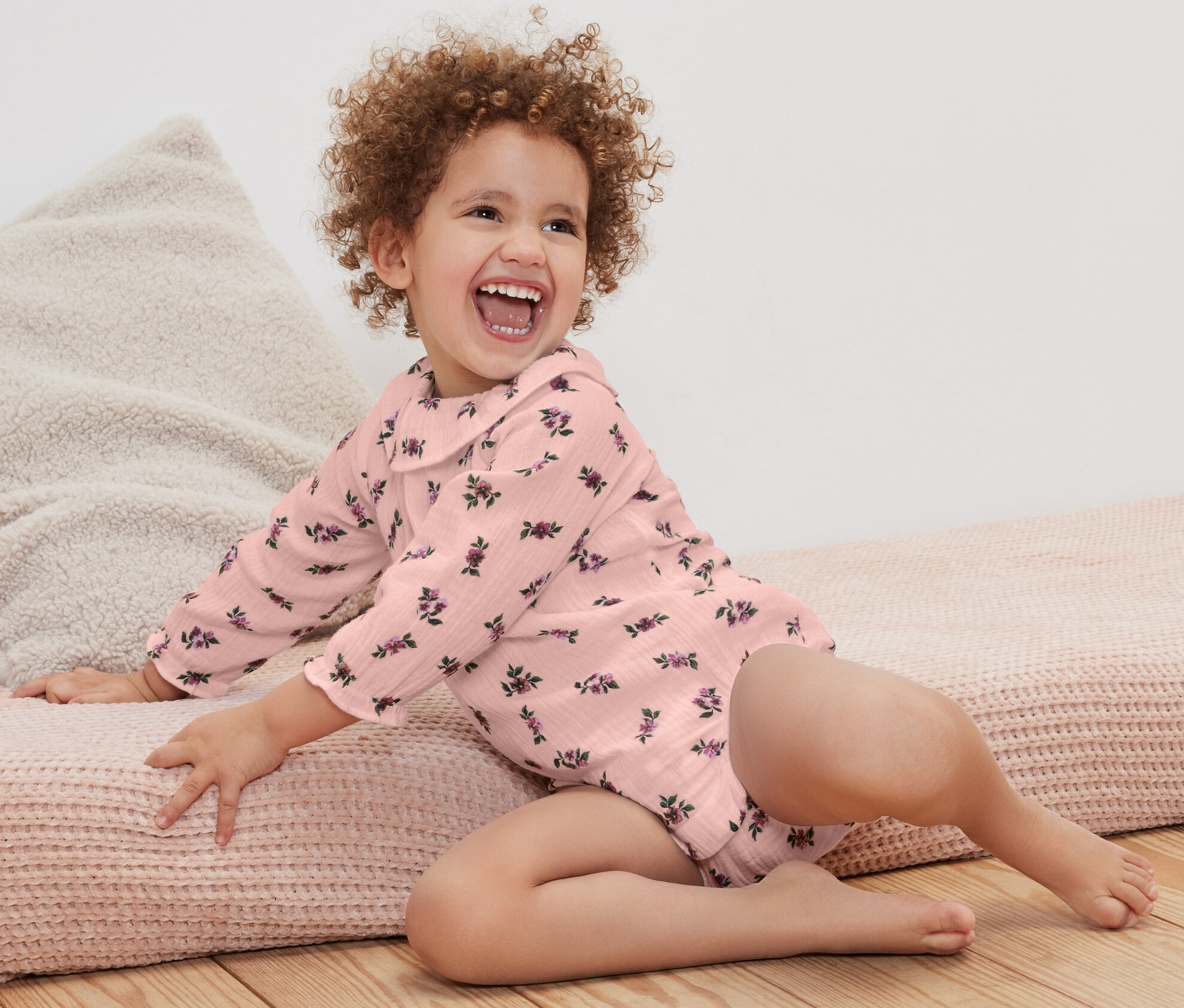 Une petite fille bouclée et souriante est assise sur un tapis et porte un body en mousseline pour bébé à motifs floraux.