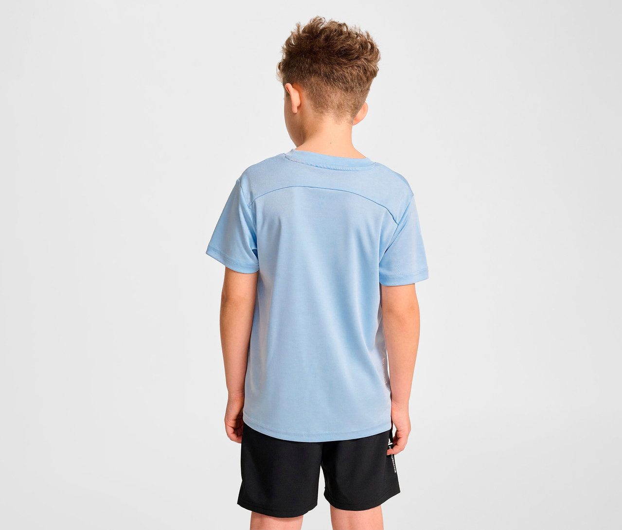 Garçon vu de dos portant un t-shirt d’entraînement HUMMEL HMLJR pulse bleu clair et un short noir.