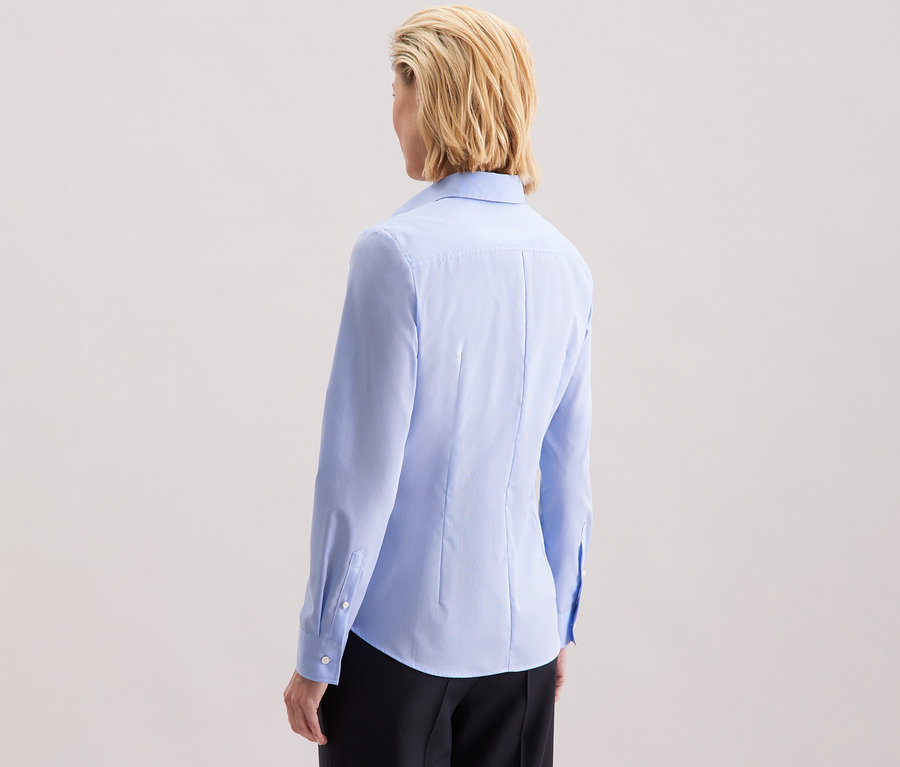 Vue arrière d'une femme aux cheveux blonds mi-longs portant un chemisier bleu clair Slim Fit non iron Seidensticker.