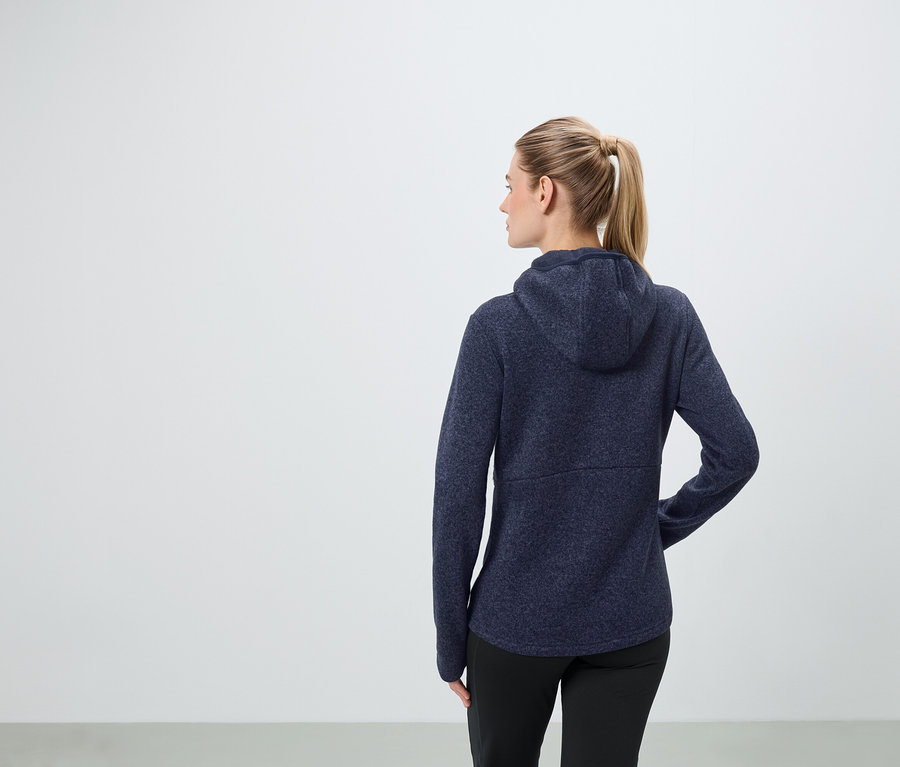 Une femme aux cheveux blonds attachés en queue de cheval porte un sweat à capuche bleu marine et un pantalon noir.