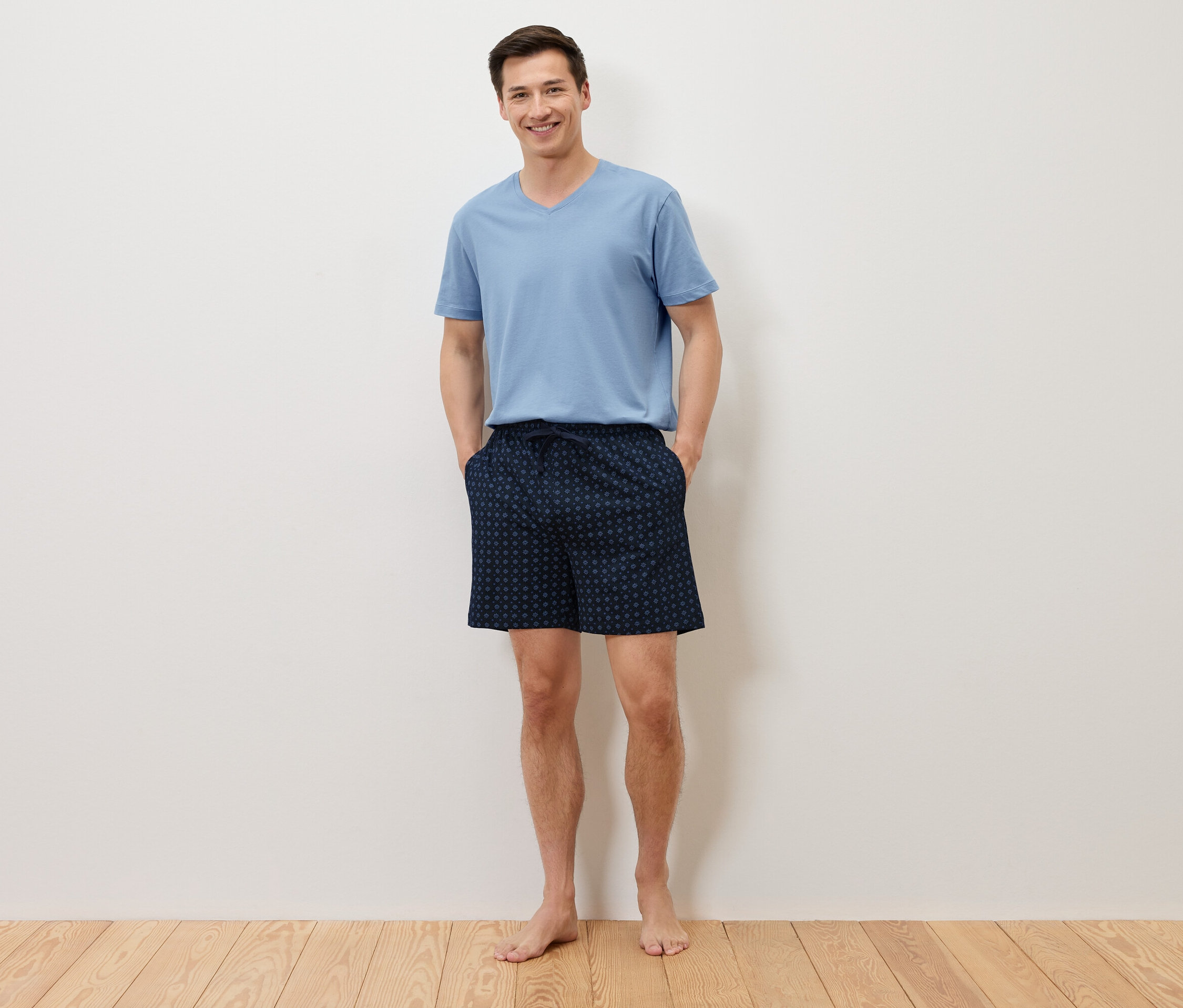 Un homme se tient pieds nus, les mains dans les poches de son short bleu marine à pois, et porte un t-shirt bleu clair devant un mur blanc.