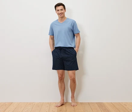 Un homme se tient pieds nus, les mains dans les poches de son short bleu marine à pois, et porte un t-shirt bleu clair devant un mur blanc.