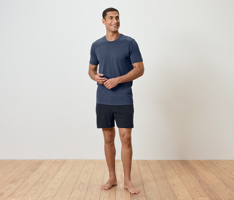 Un homme se tient pieds nus sur un plancher en bois, portant un t-shirt bleu et un short noir.