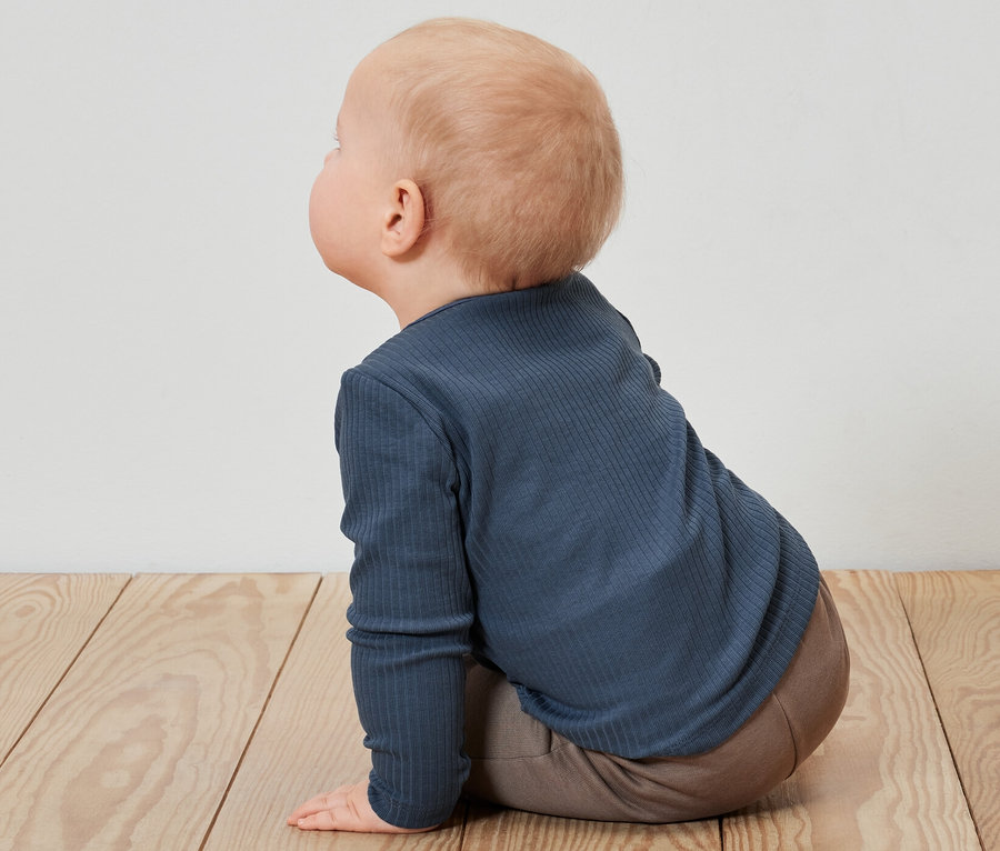 Un bébé est assis sur un plancher en bois et porte un t-shirt bleu à manches longues avec une structure côtelée.