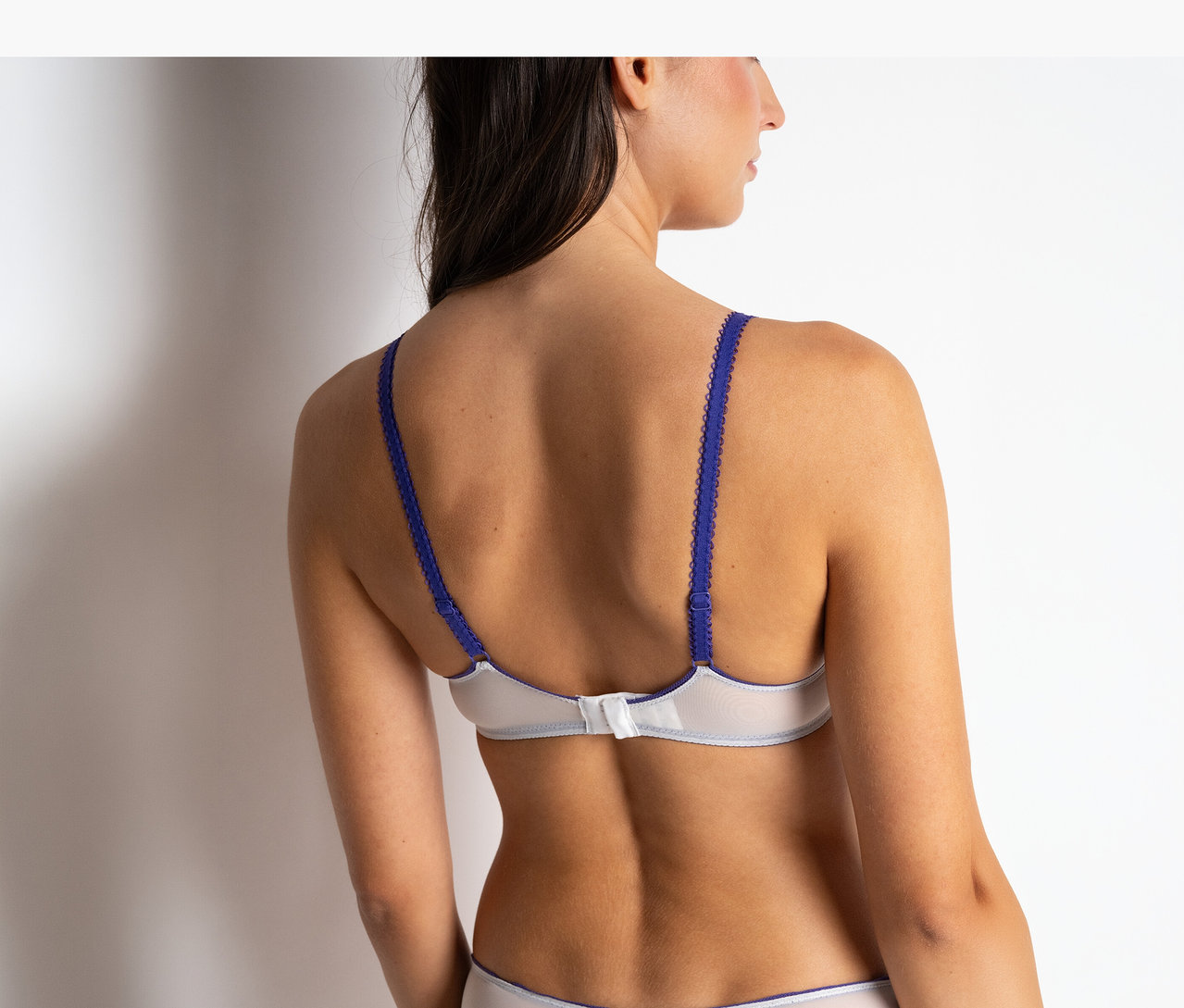 Vue de dos d'une femme portant un soutien-gorge plongeant à armatures « Chloe » Passionata blanc à bretelles bleues et une culotte assortie.