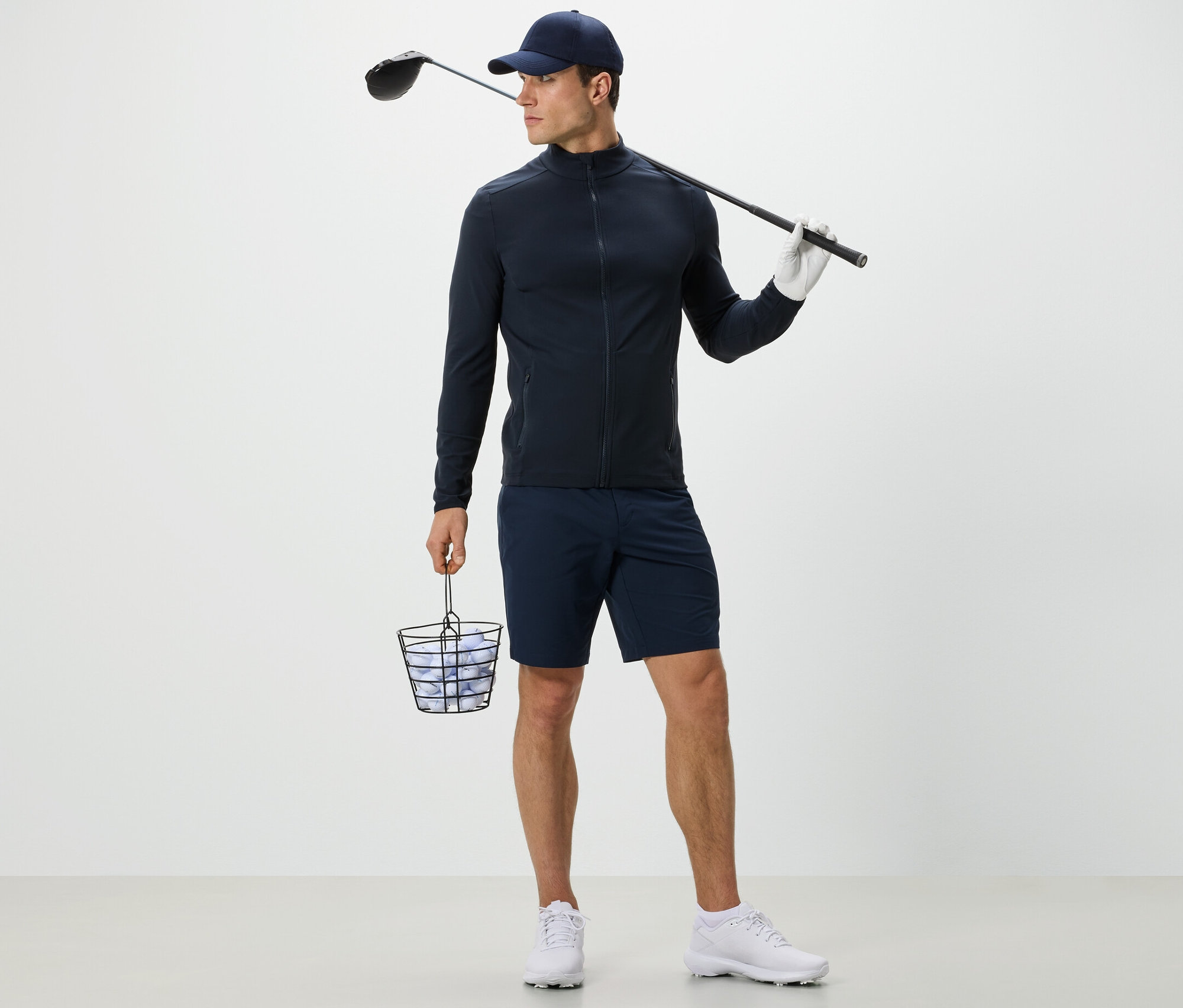 Un homme portant une veste fonctionnelle bleu marine tient un panier de balles de golf et un club de golf.
