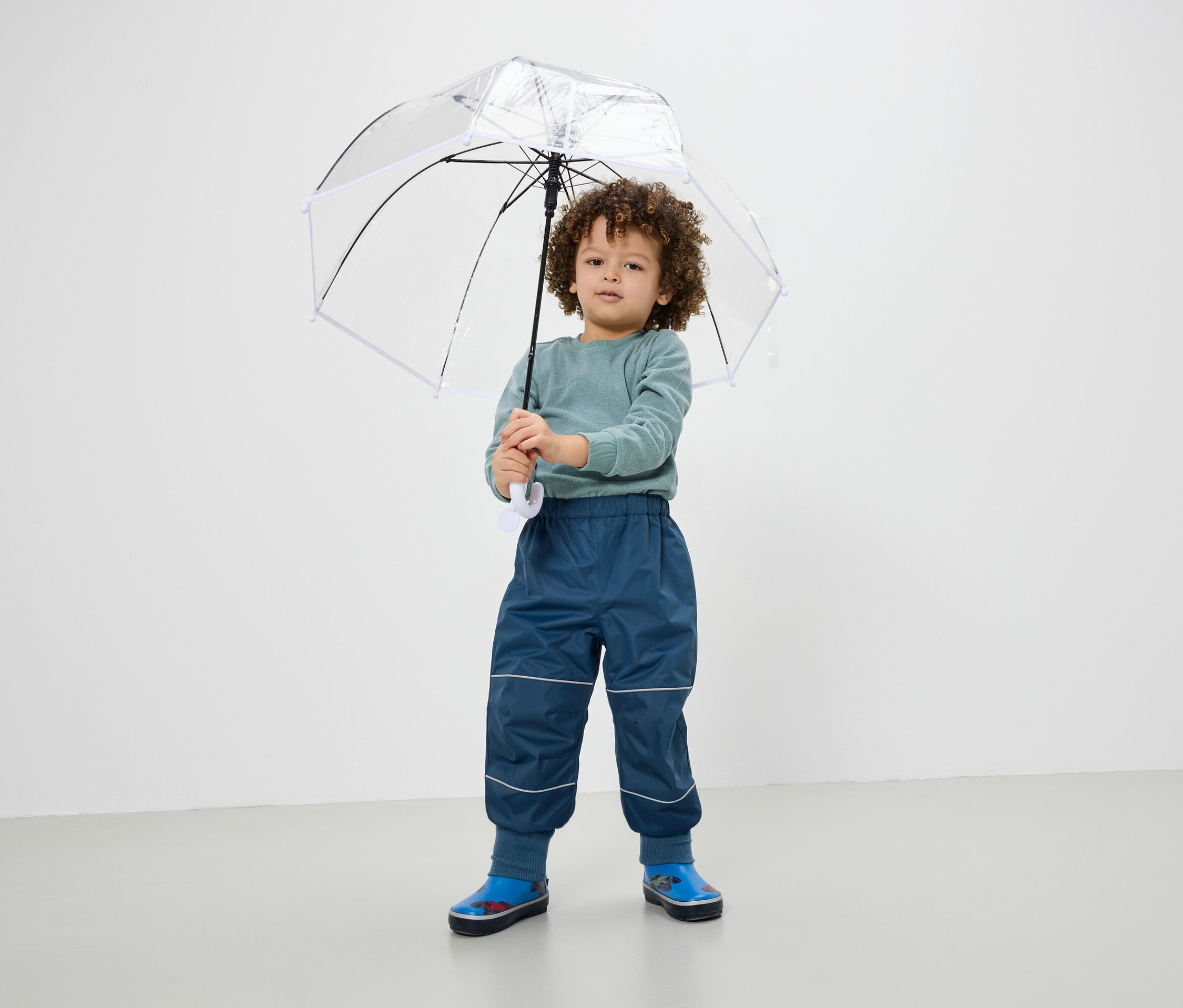 Un garçon tient un parapluie transparent, porte un pantalon bleu et des bottes en caoutchouc.