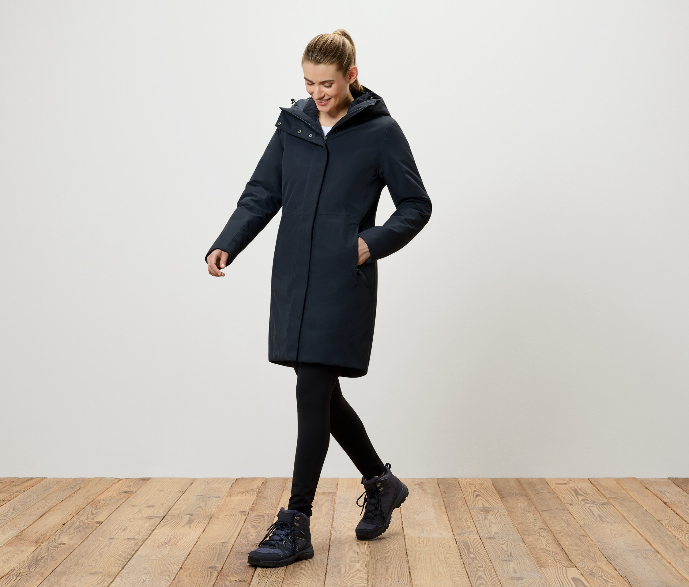 Une femme marche sur un plancher en bois et porte un manteau bleu foncé à capuche, des leggings noirs et des chaussures bleu foncé.