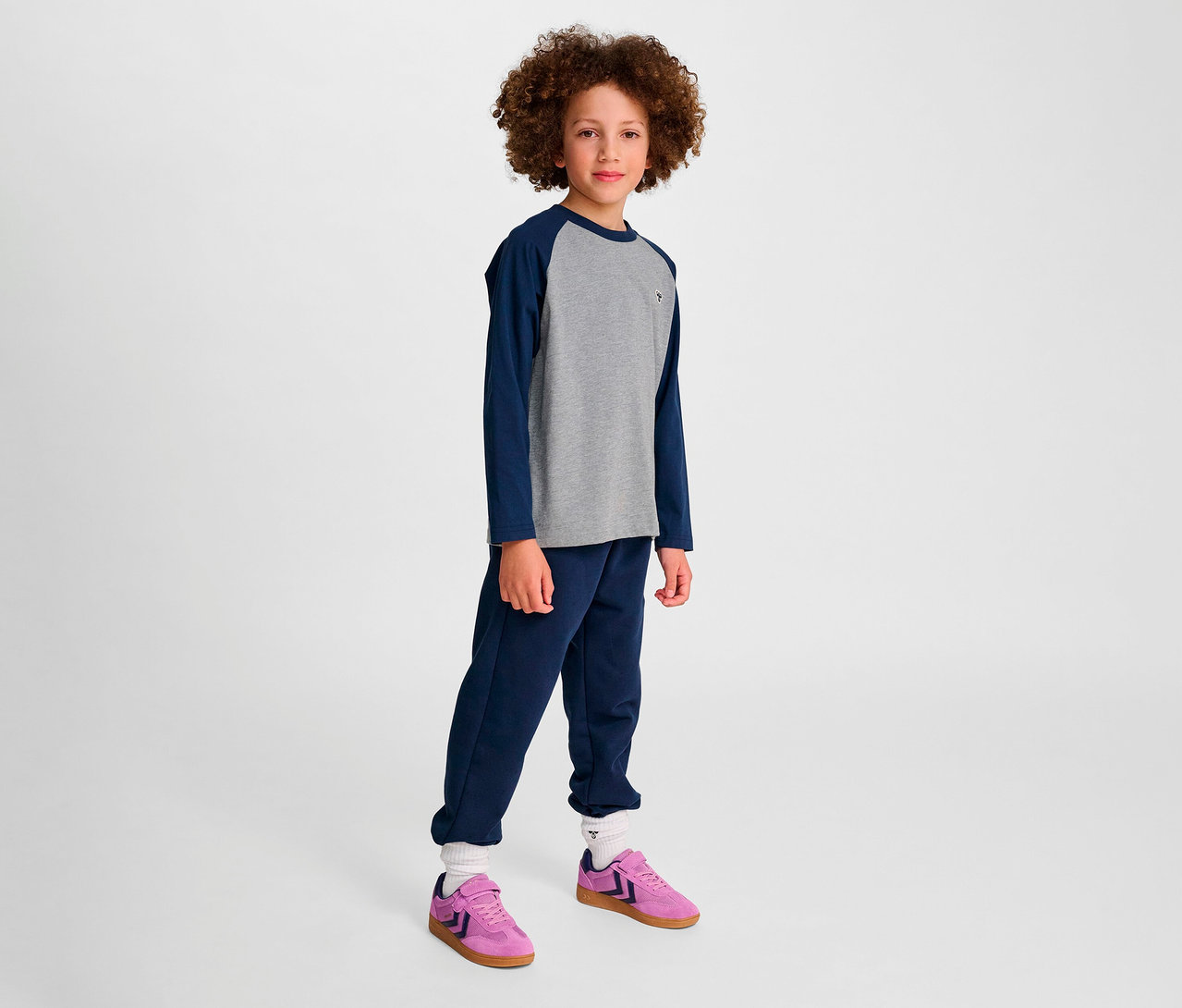 Un garçon debout porte un pantalon de survêtement bleu et un t-shirt ample à manches raglan HUMMEL HMLJR BEE bleu et des chaussures roses.