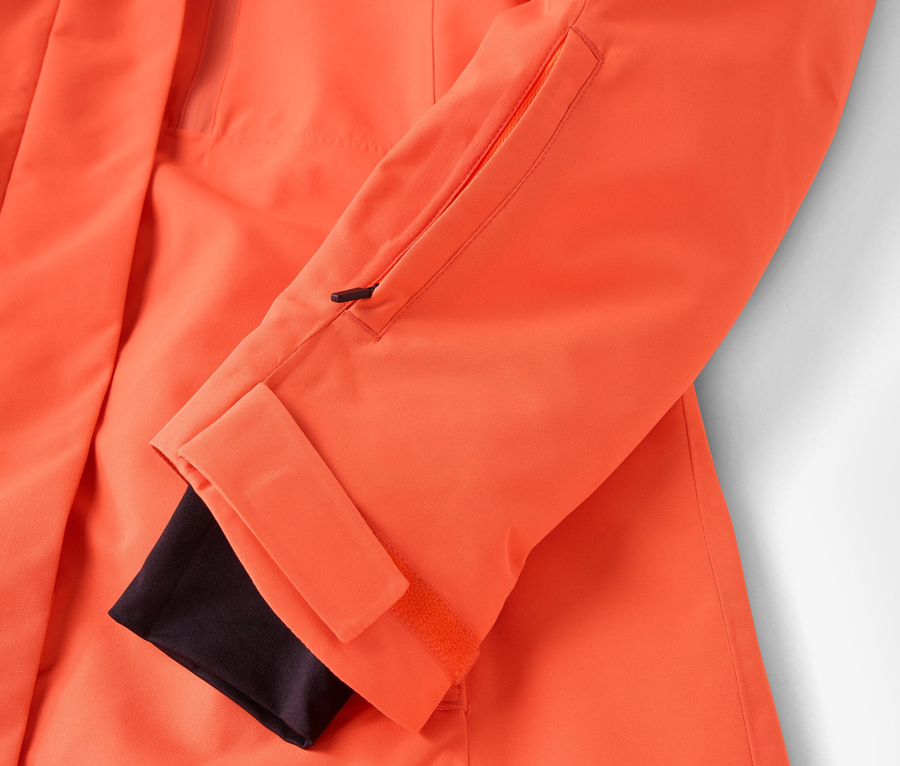 Détail d'une veste orange avec un poignet noir et une poche zippée.