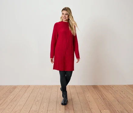 Blonde portant une robe en maille rouge, des leggings noirs et des bottes noires, debout sur un plancher en bois.