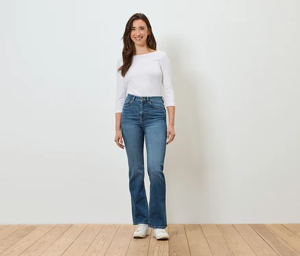 Une femme se tient devant un mur blanc et porte un jean bleu, une chemise blanche et des baskets blanches.