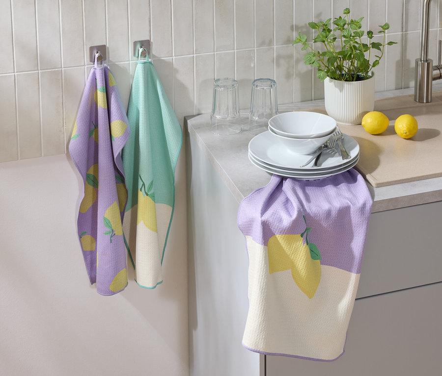 Un torchon de cuisine violet et vert avec un motif de citron est accroché au mur. Des assiettes, un bol, des verres, des couverts, un citron et une fleur sont posés sur le plan de travail.