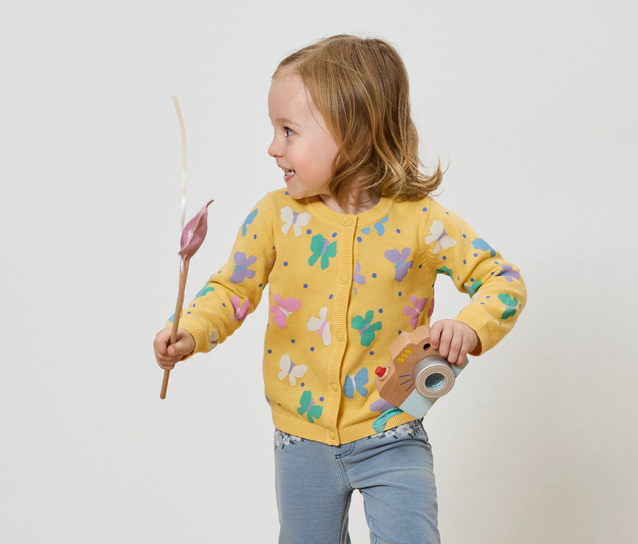 Fillette en gilet jaune à papillons tenant un appareil photo jouet et une fleur.