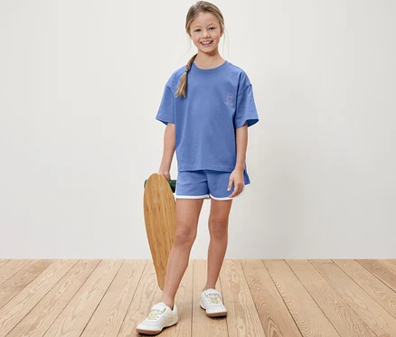 Une fille tient un skateboard, porte un t-shirt bleu et un short bleu.