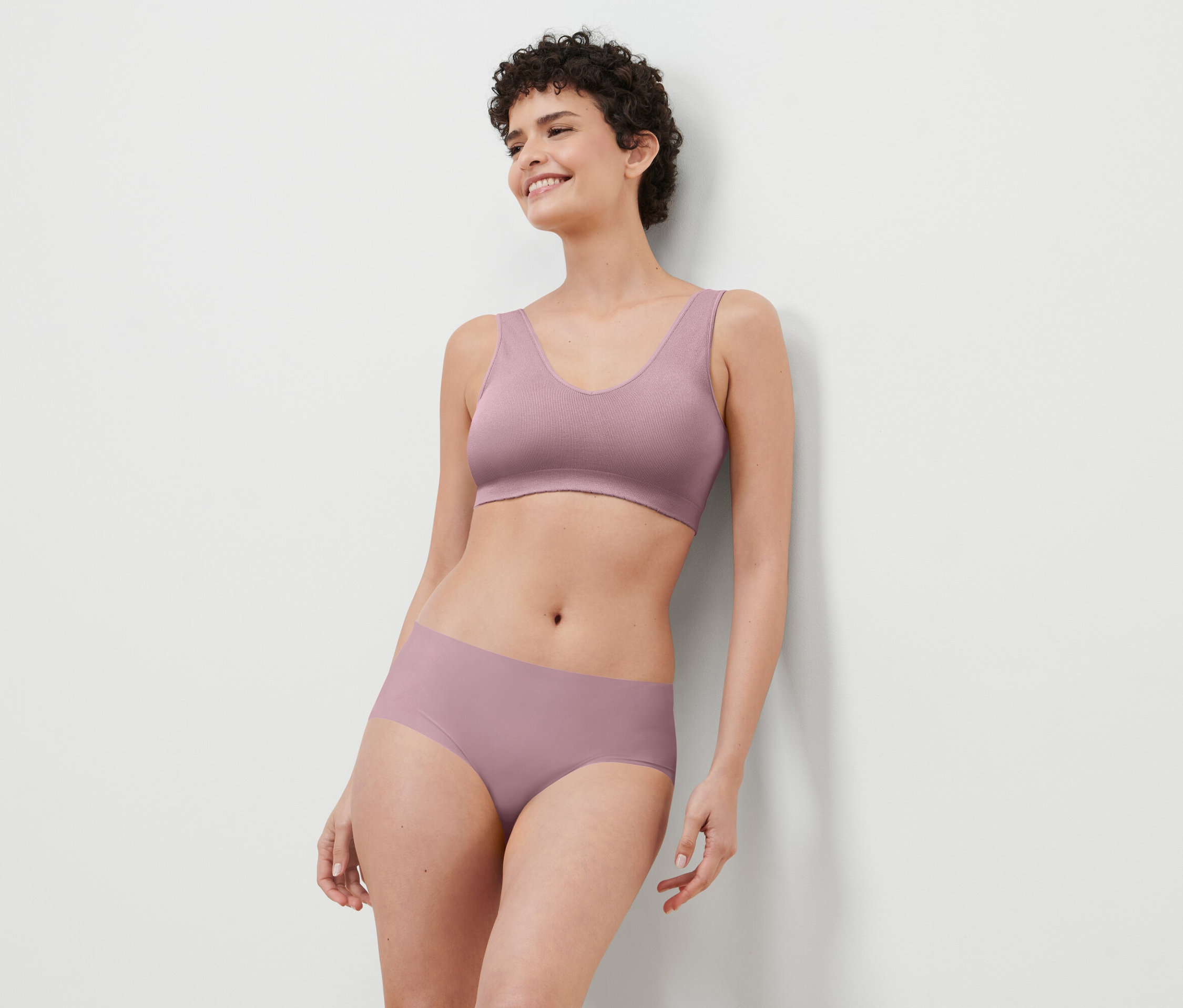 Femme souriante aux cheveux courts, appuyée contre un mur, portant un soutien-gorge et une culotte lilas.